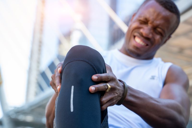 El corredor afroamericano se dobla agarrándose la rodilla mientras sufre un dolor intenso por una lesión aguda en la rodilla, los atletas se lastiman las rodillas.