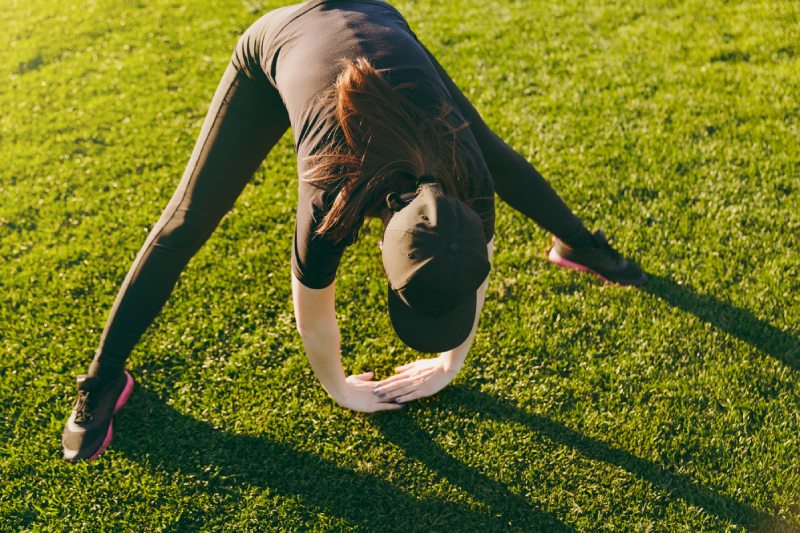 Joven atlética con uniforme negro, gorra haciendo ejercicios deportivos, calentamiento, estiramiento antes de correr en el césped verde en el parque del campo de golf al aire libre en el soleado día de verano.  Fitness, concepto de estilo de vida saludable