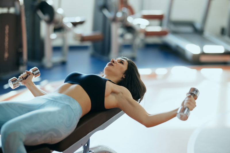 Una atractiva mujer culturista está levantando pesas mientras se acuesta en el banco del gimnasio.